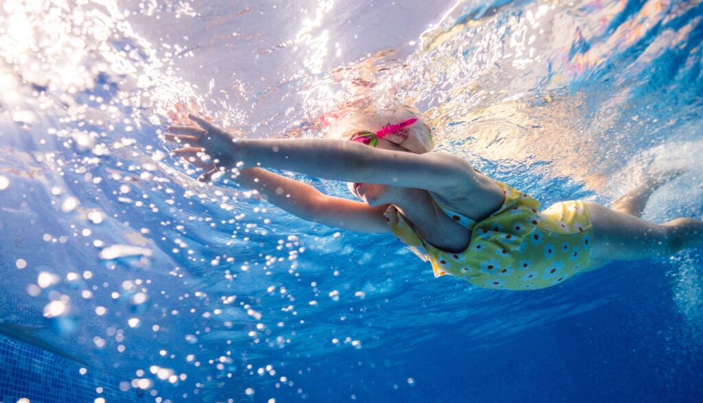 Kind mit gelbem Badeanzug und Schwimmbrille taucht unter Wasser in einem Schwimmbecken